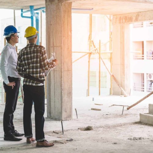Two construction workers wearing hard hats, one blue and one yellow, stand inside a building under construction, looking at blueprints. Sunlight streams in, creating a warm atmosphere.