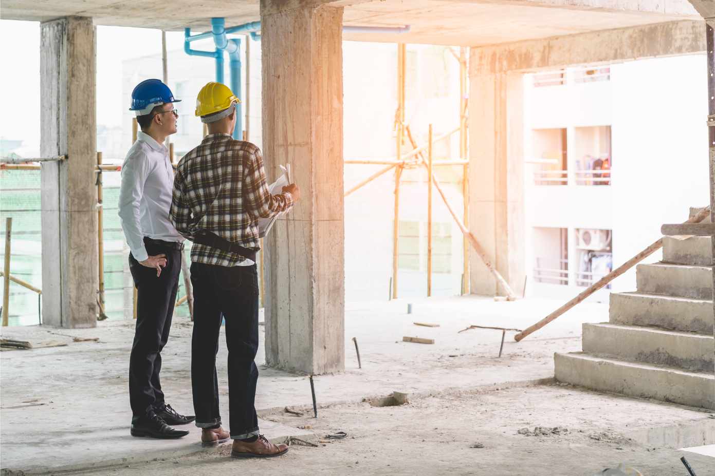 Two construction workers wearing hard hats, one blue and one yellow, stand inside a building under construction, looking at blueprints. Sunlight streams in, creating a warm atmosphere.