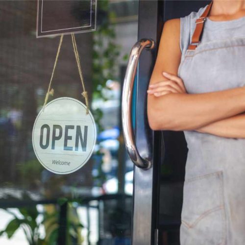 A person in an apron stands with arms crossed beside a glass door. An "OPEN" sign hangs beside them. The scene is inviting and welcoming.