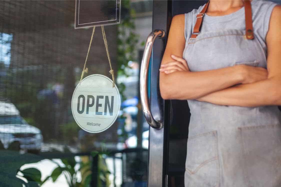A person in an apron stands with arms crossed beside a glass door. An "OPEN" sign hangs beside them. The scene is inviting and welcoming.