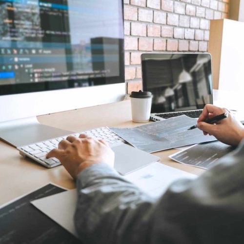 A person works at a desk with dual monitors displaying code, holding a pen over documents. A coffee cup sits nearby. The atmosphere is focused and tech-driven.