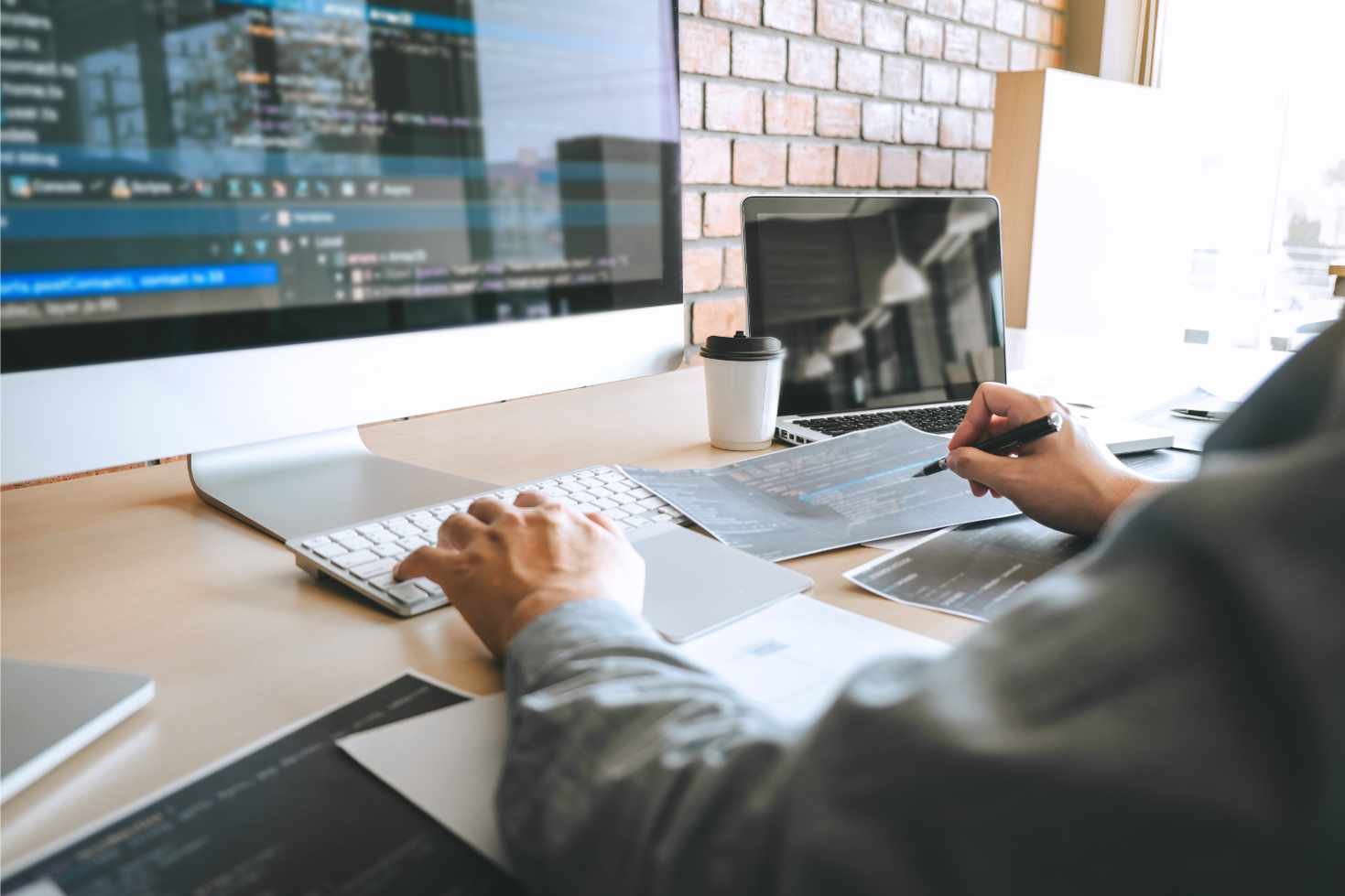 A person works at a desk with dual monitors displaying code, holding a pen over documents. A coffee cup sits nearby. The atmosphere is focused and tech-driven.